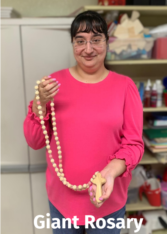 Woman in pink holds a giant wooden rosary. Smiles at the camera.