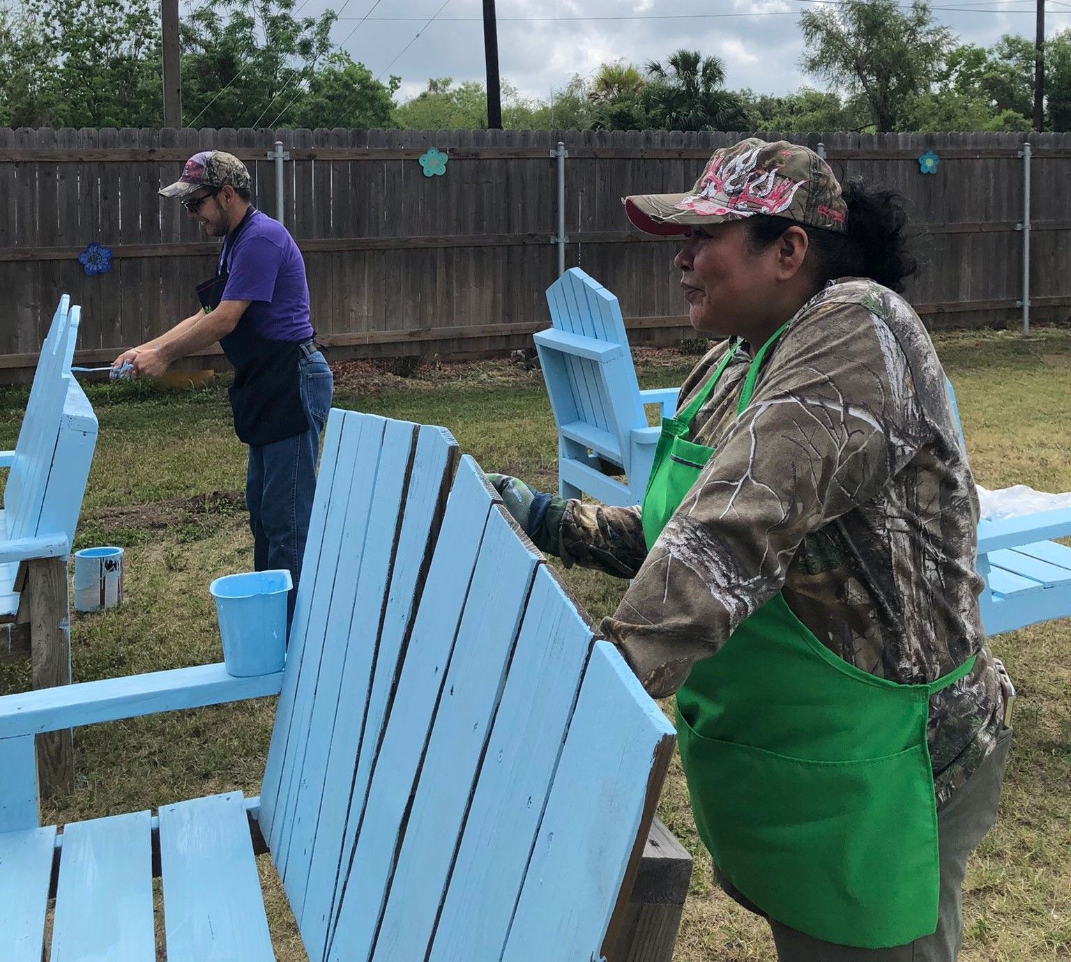 A man and a woman are painting blue chairs in a yard.