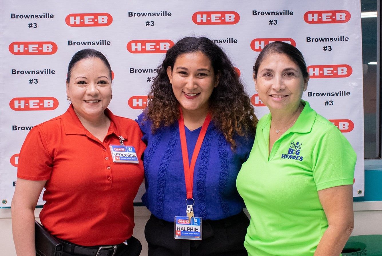 Three women are posing for a picture in front of a h-e-b sign.