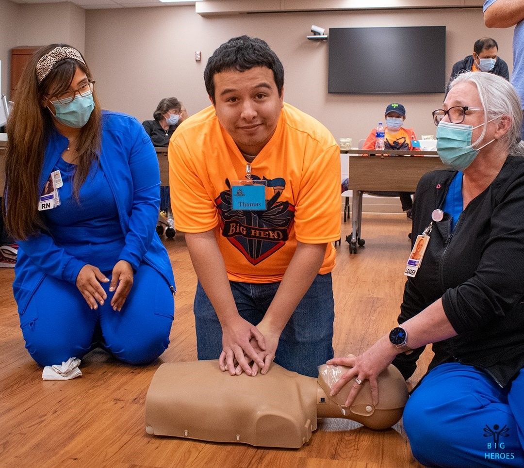 A group of people wearing masks are kneeling on the floor while a man performs a heart massage on a mannequin.