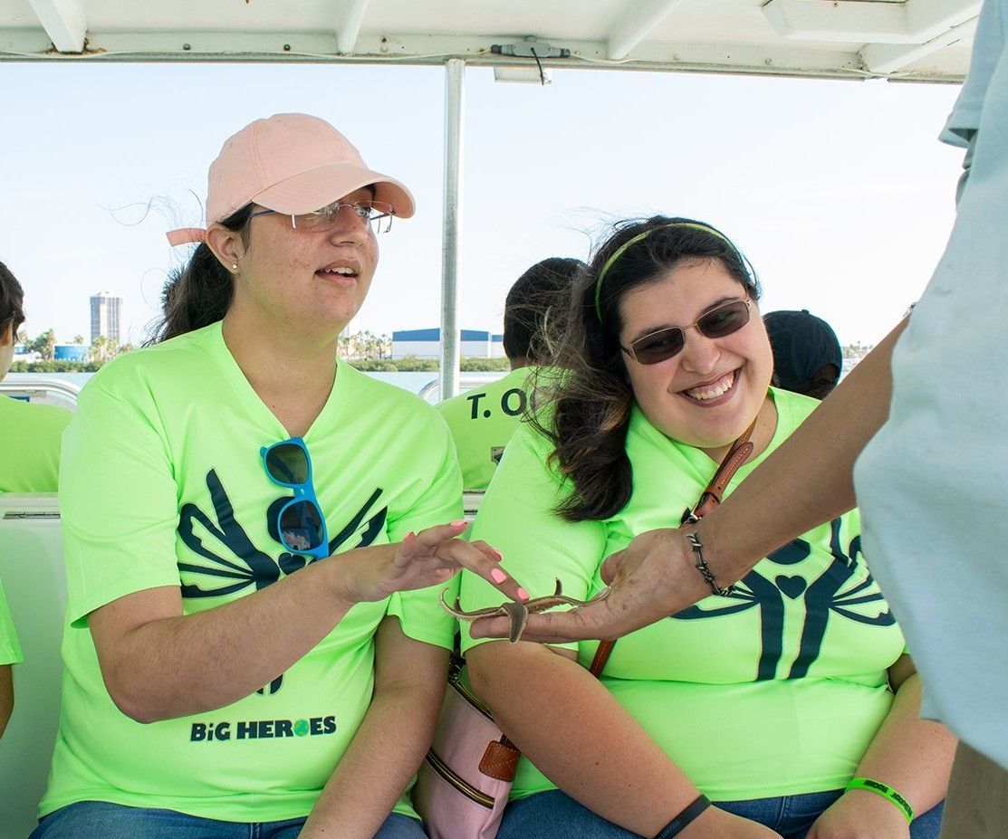 Two women wearing green shirts that say big heroes are sitting on a boat