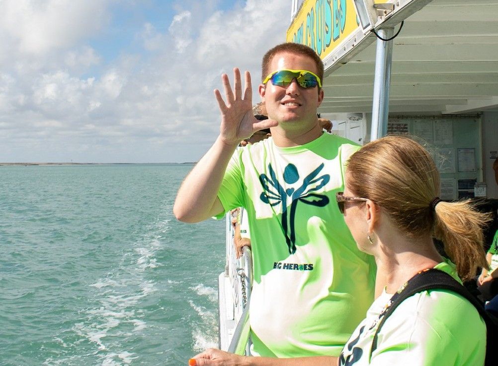 A man in a green shirt with a dragonfly on it waves to a woman on a boat