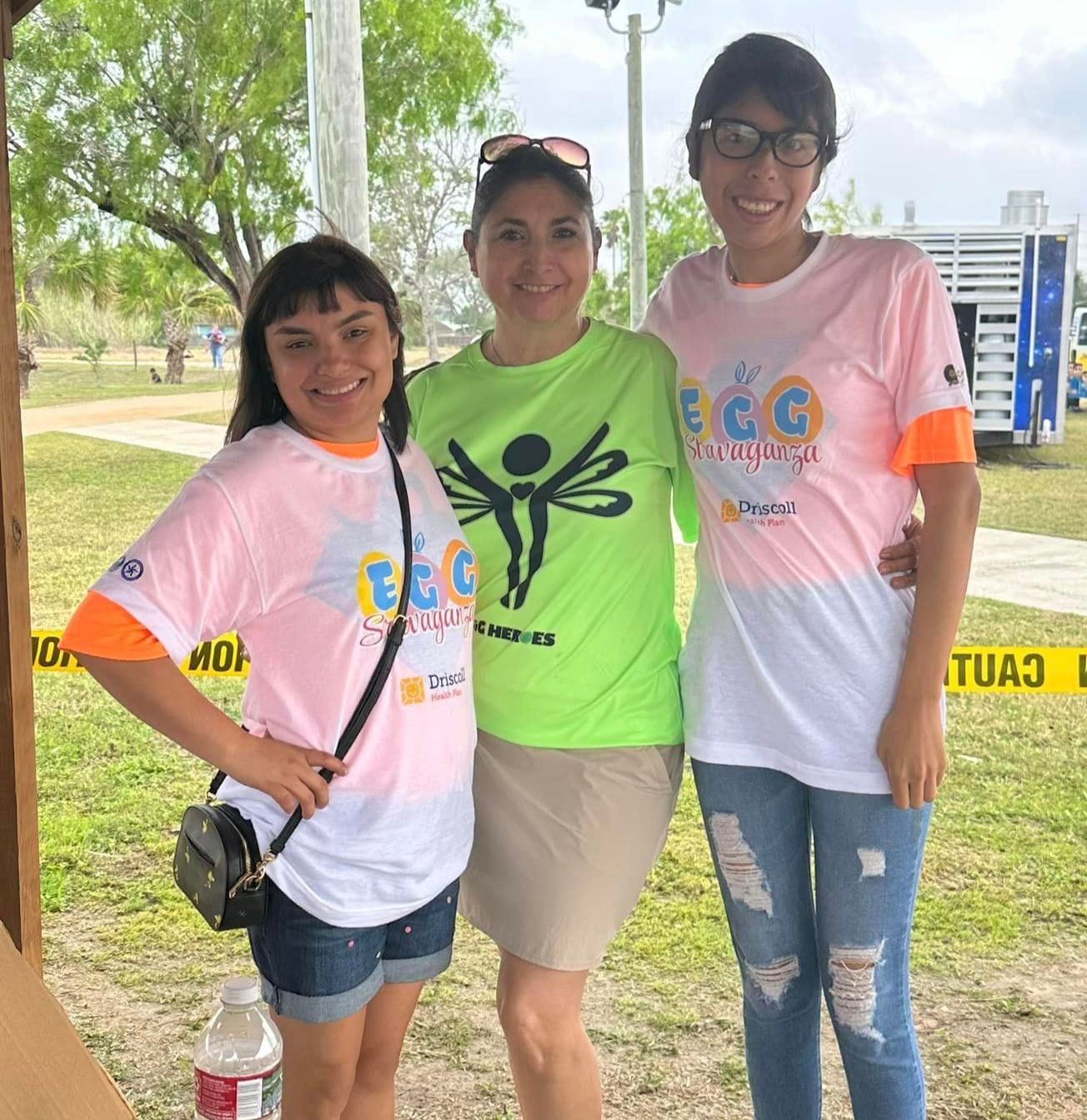 Three women are posing for a picture with one wearing a green shirt with a dragonfly on it