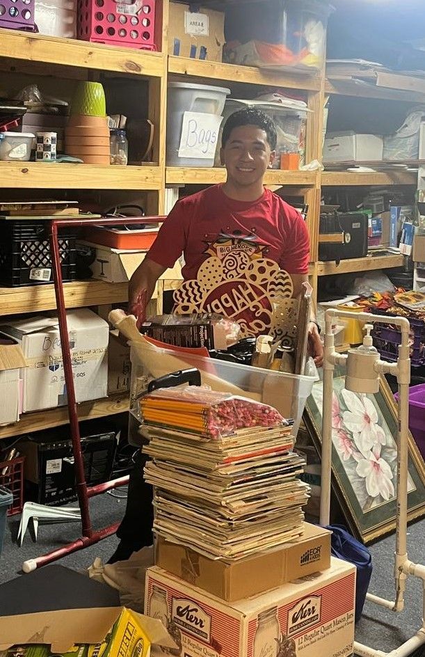 A man in a red shirt is standing in a cluttered room.