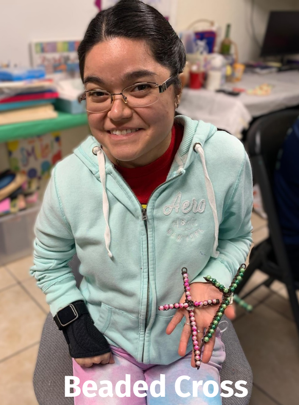 Woman smiling, holding beaded crosses. Wearing glasses, teal jacket, and has a brace on her left hand.