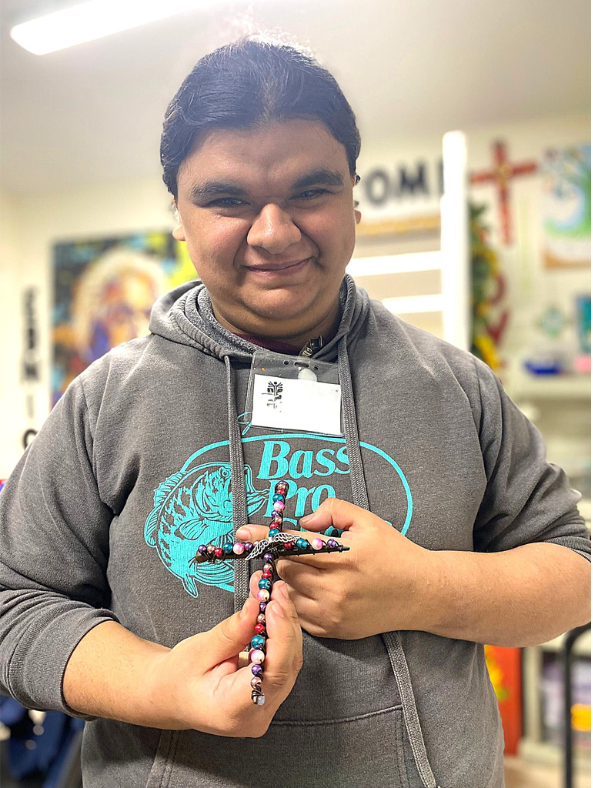 A smiling person in a grey hoodie holds a beaded cross indoors.