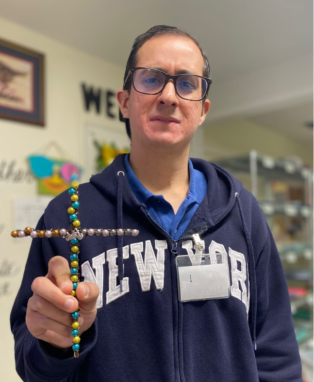 Man in glasses holding a beaded cross, wearing a blue sweatshirt, standing inside a room with