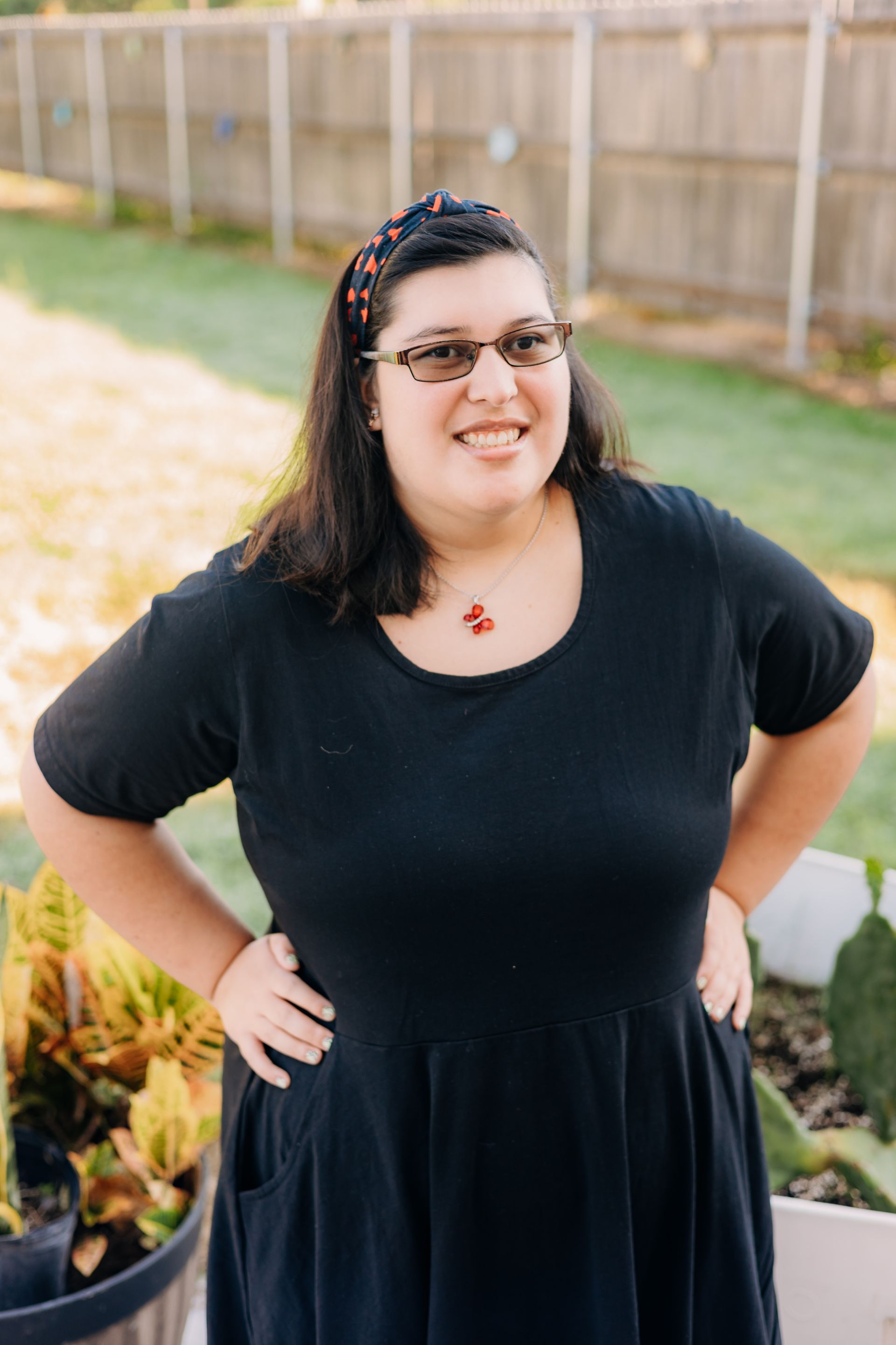 A woman in a black dress and glasses is standing in front of a fence.