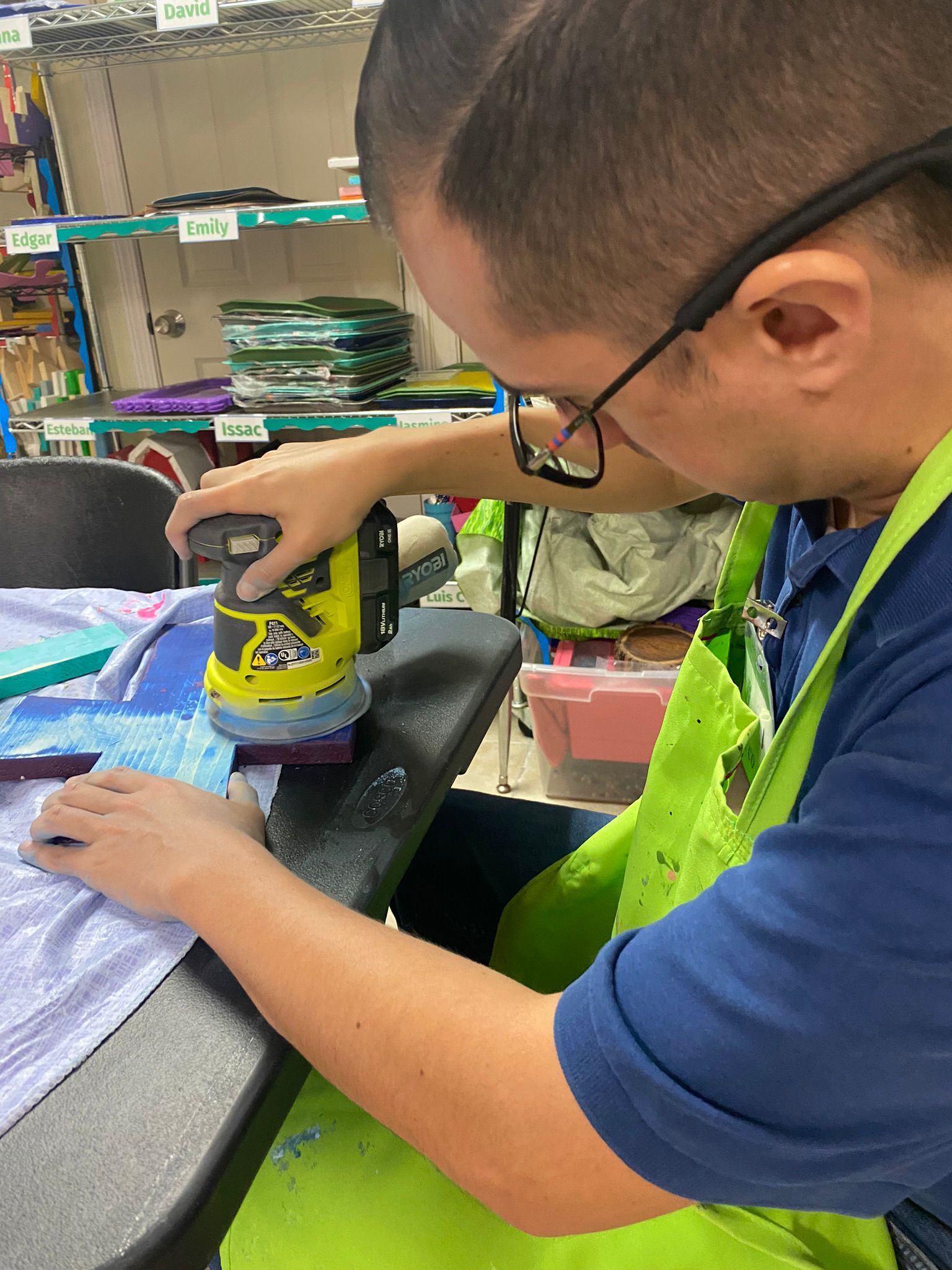 A man in a green apron is using a sander on a piece of wood.