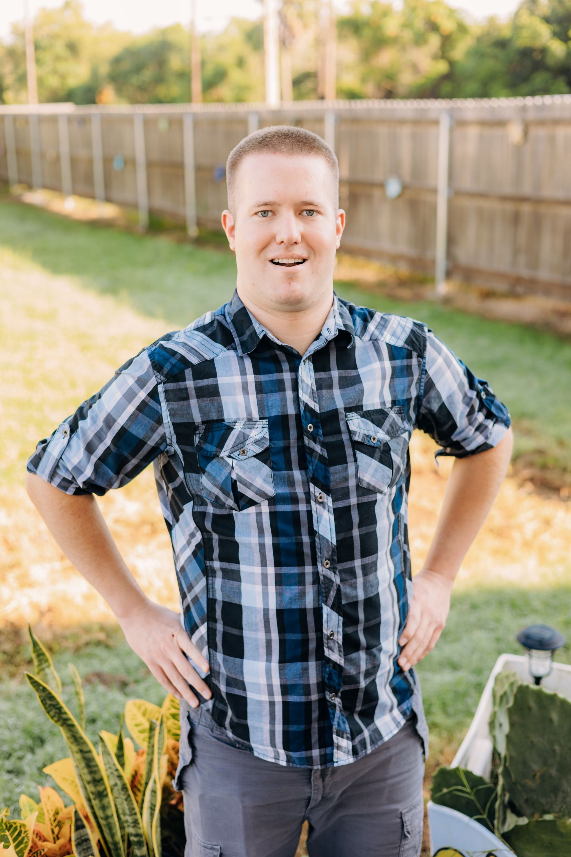 A man in a plaid shirt is standing in a yard with his hands on his hips.