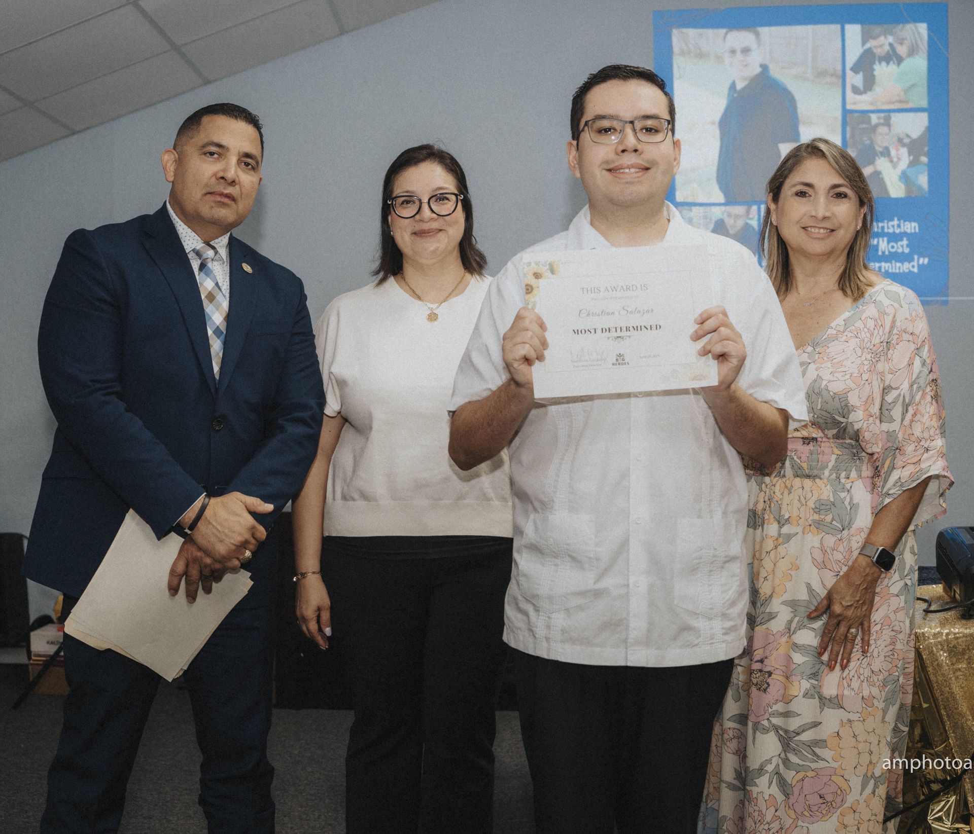 Four people pose; a man holds a certificate, smiling, flanked by two women and a man in a suit, indoors.