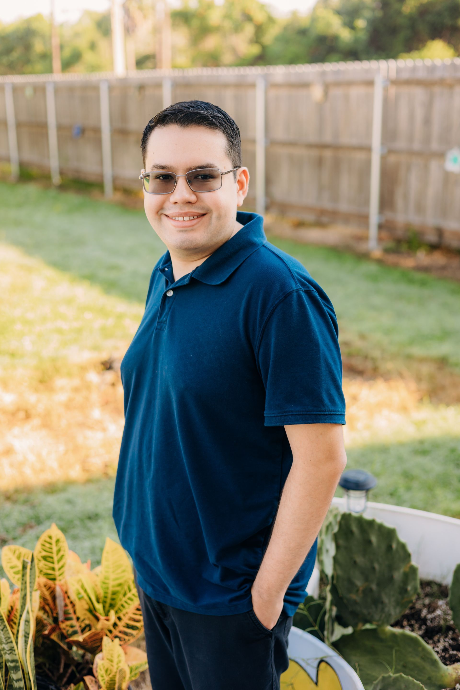 A man wearing sunglasses and a blue shirt is standing in a garden with his hands in his pockets.