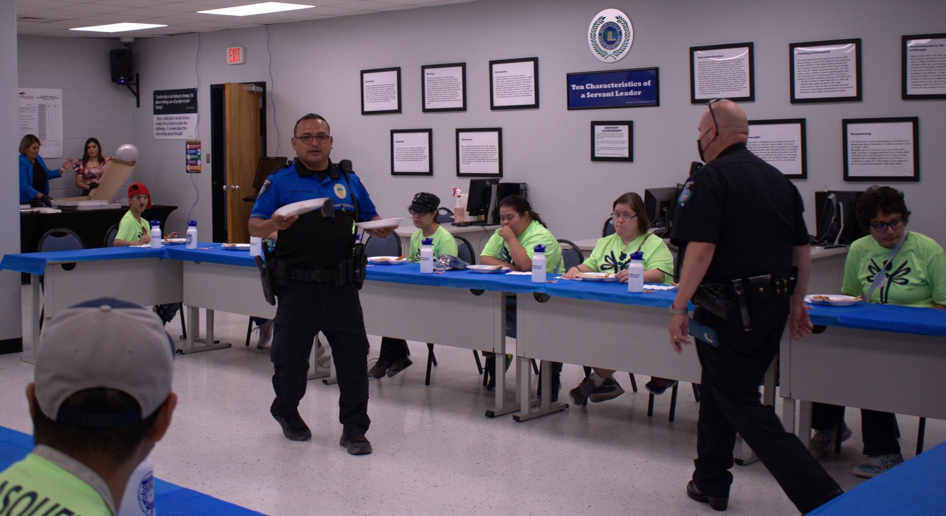 A police officer is standing in front of a group of people sitting at tables.
