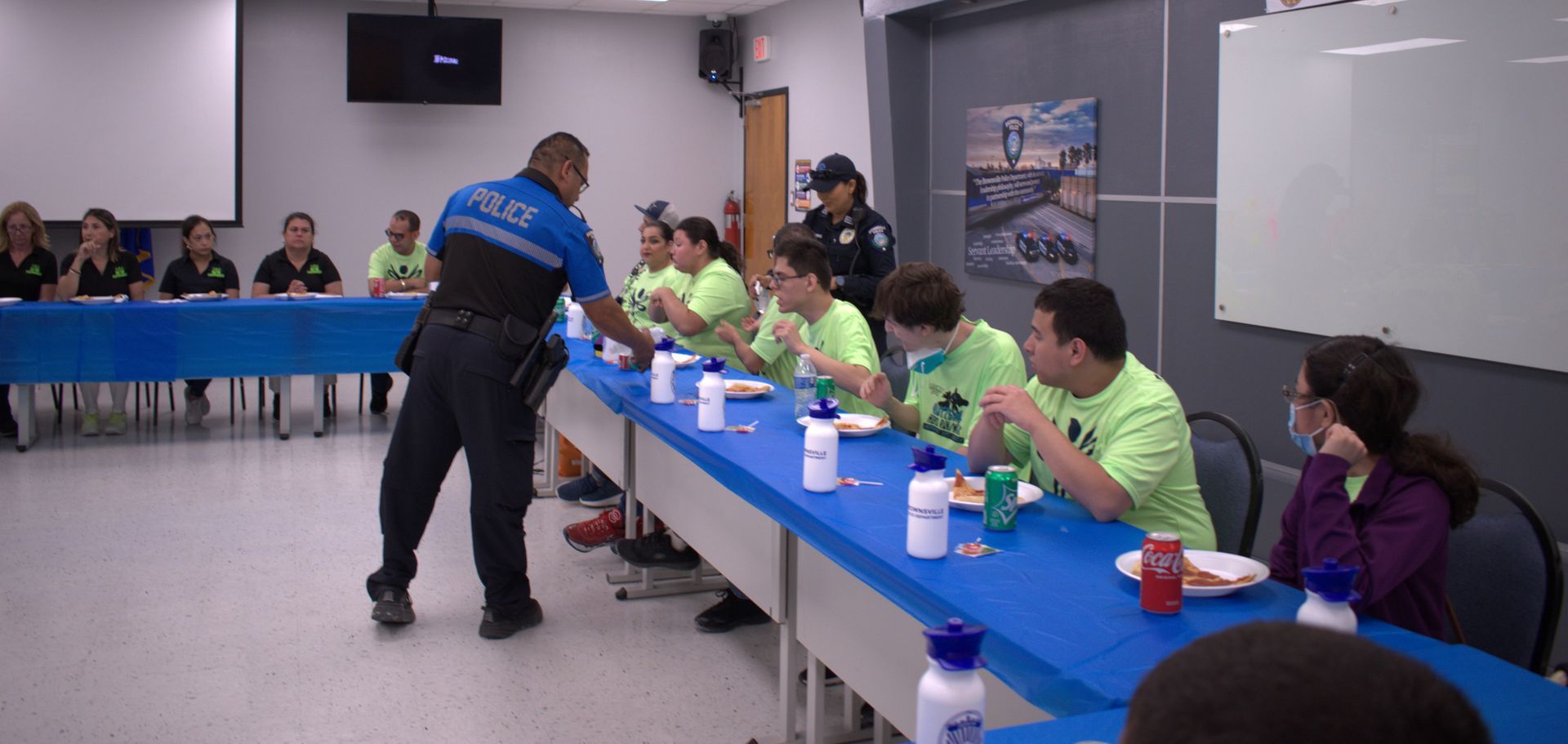 A police officer is standing in front of a long table with people sitting at it.