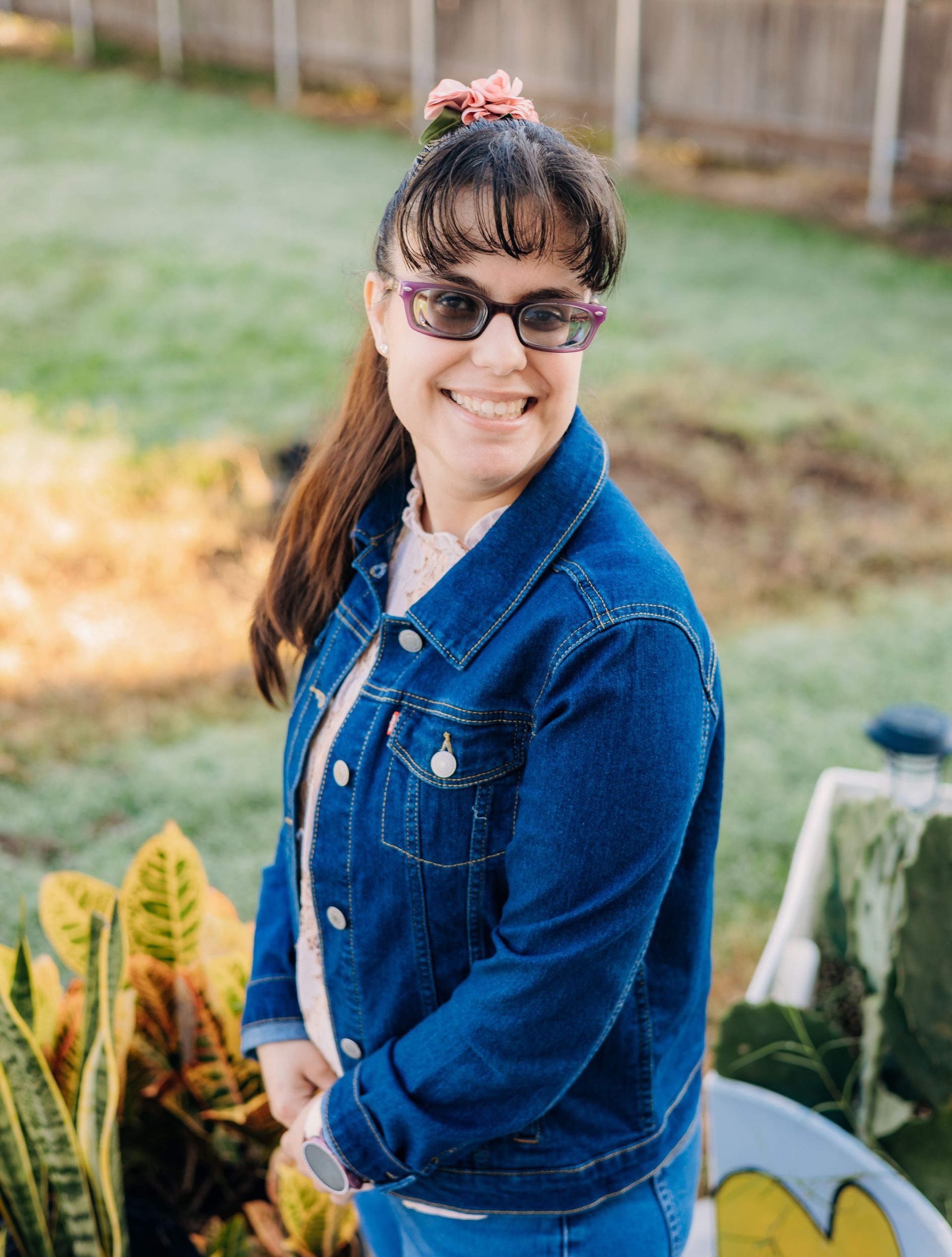 A woman wearing sunglasses and a denim jacket is standing in a garden.