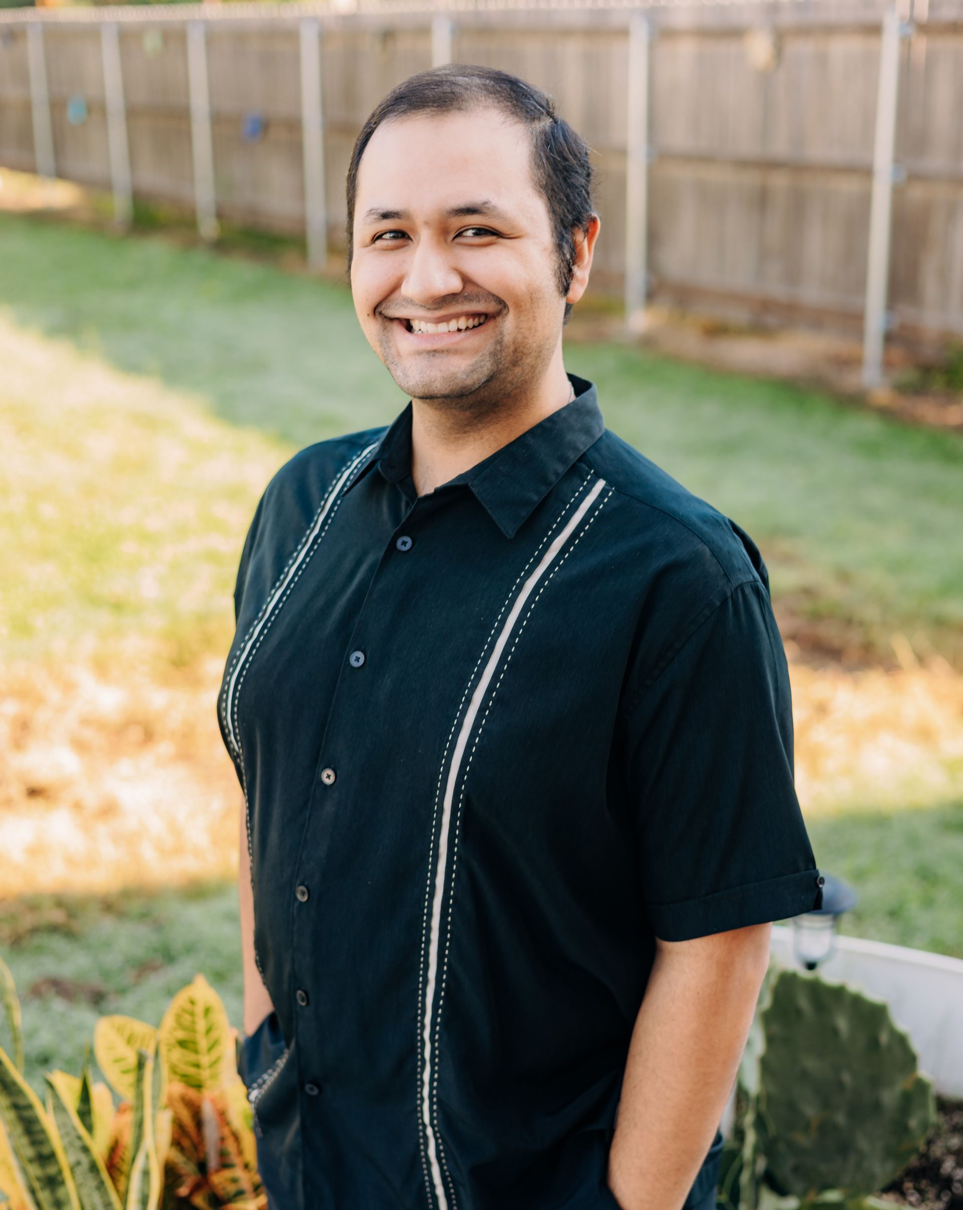 A man in a black shirt is standing in front of a fence with his hands in his pockets.