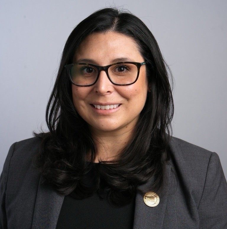A woman in a blue floral shirt is smiling in front of a bush.