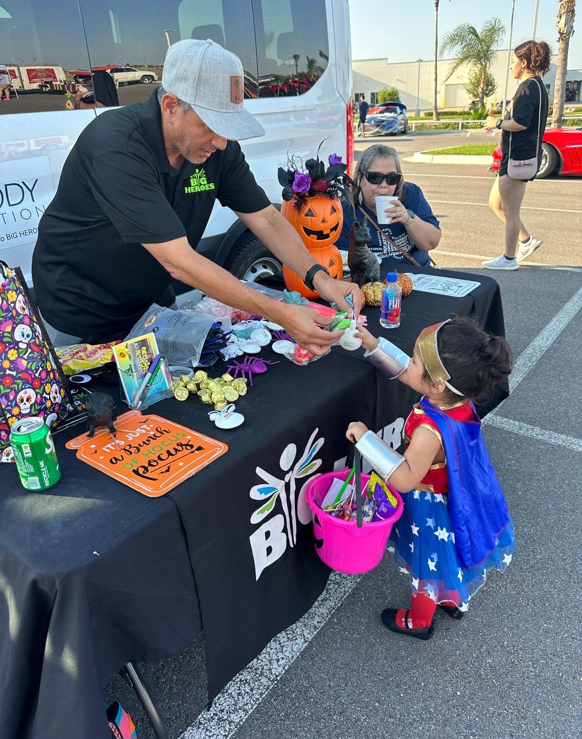 A little girl in a wonder woman costume is getting candy from a man at a table.