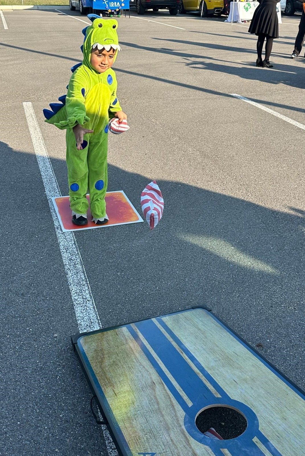 A young boy in a dinosaur costume is playing a game of cornhole in a parking lot.