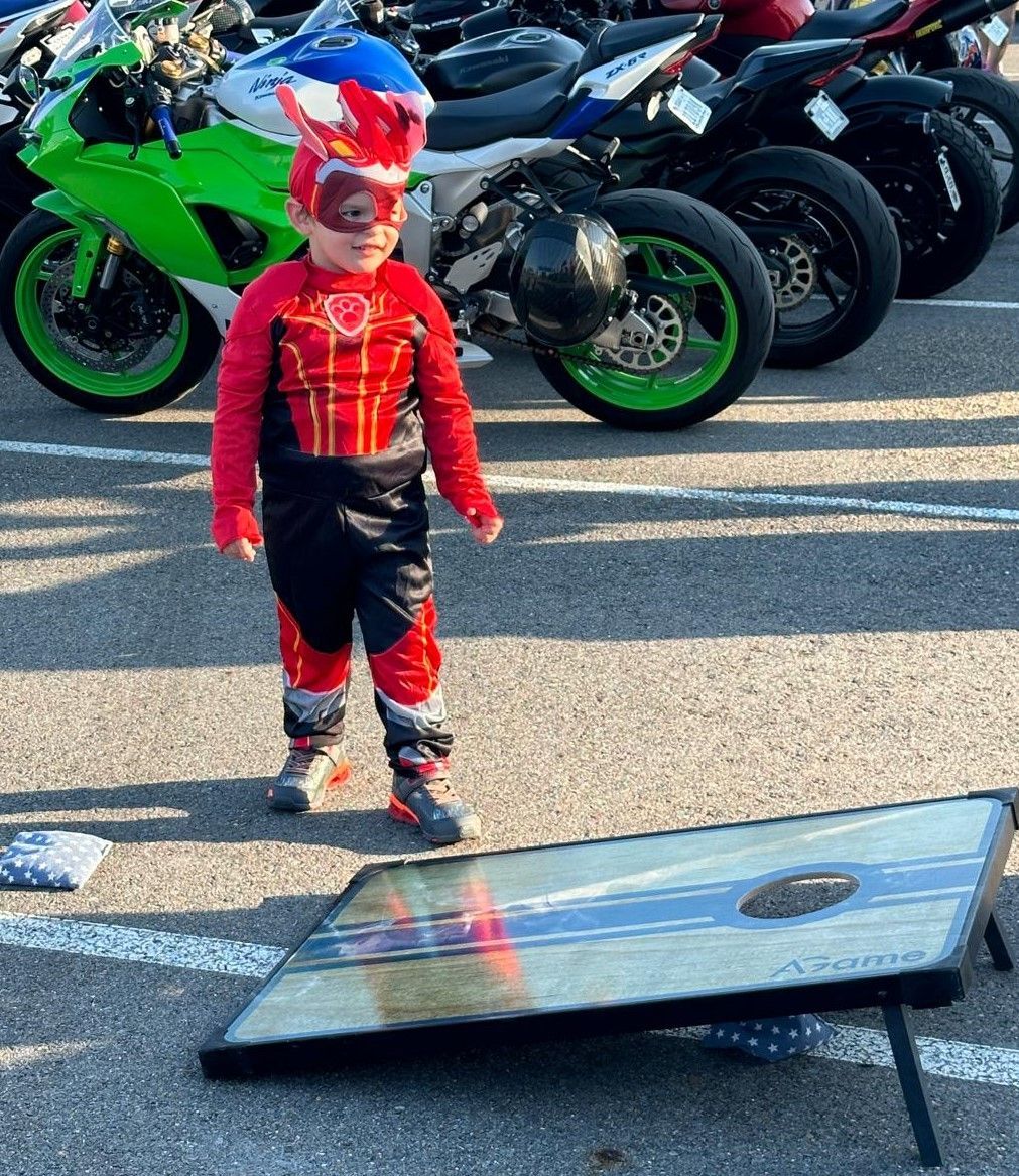 A little boy in a superhero costume is standing next to a cornhole board in a parking lot.