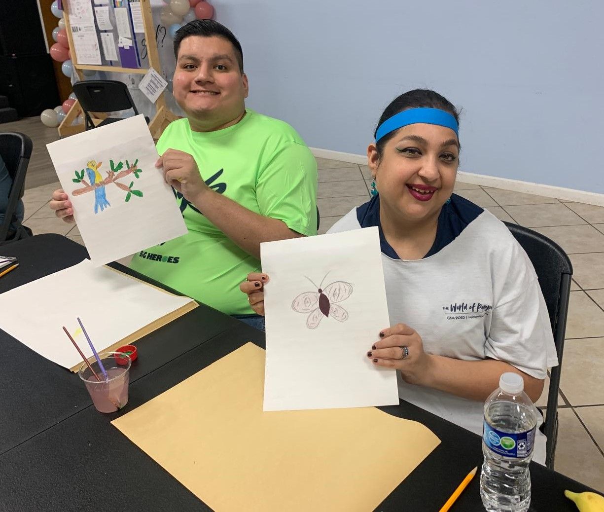 A man and a woman are sitting at a table holding drawings of a tree and a butterfly.