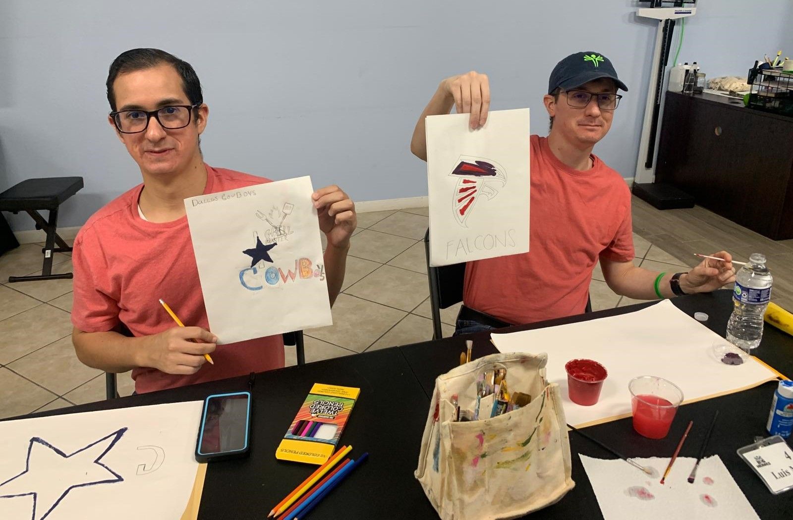 Two men are sitting at a table holding up drawings.