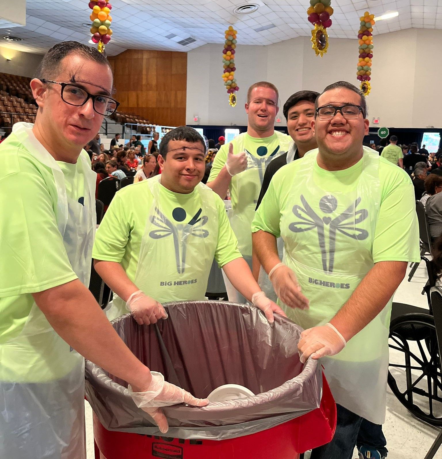 A group of men in green shirts are standing around a trash can