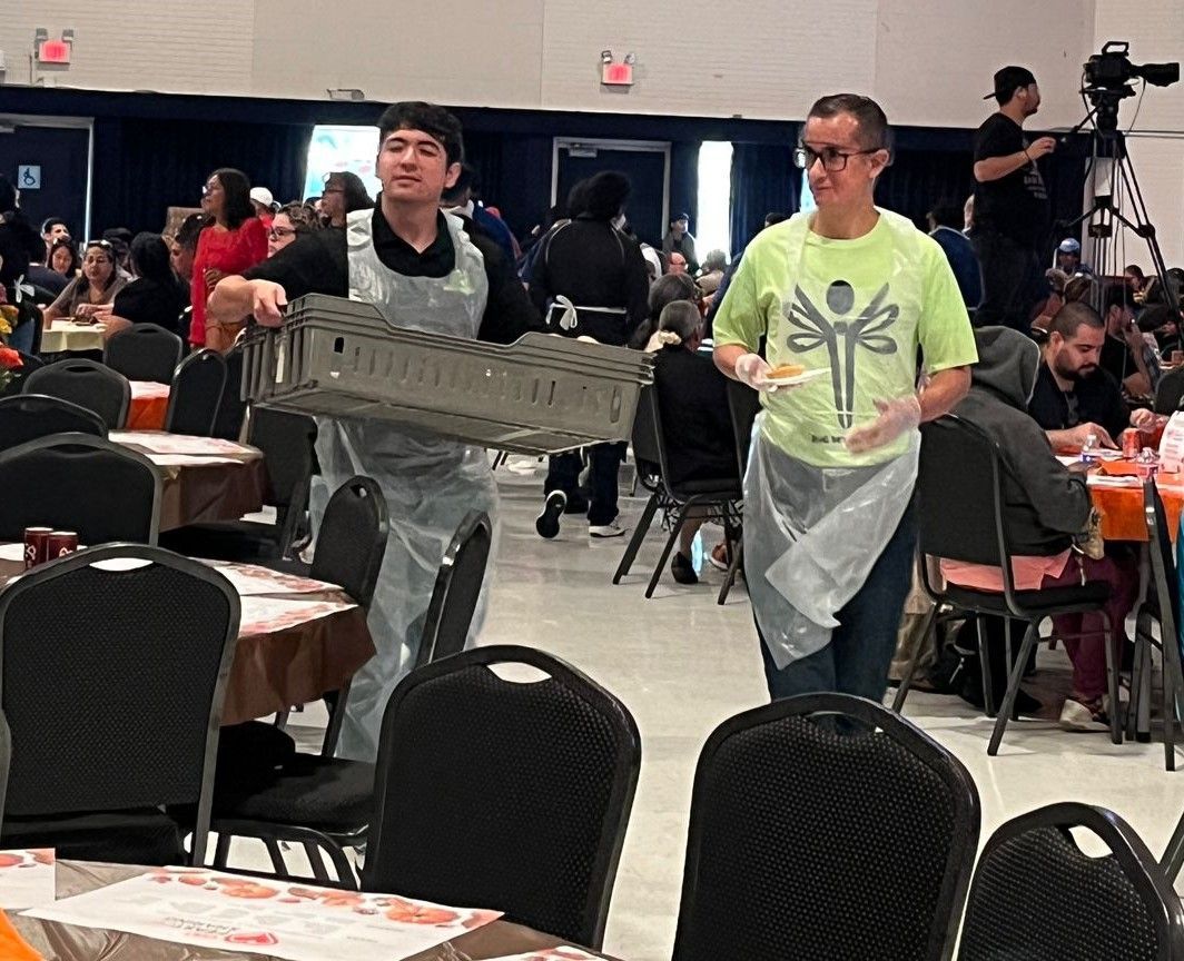 Two men are carrying a tray of food in a room with tables and chairs.