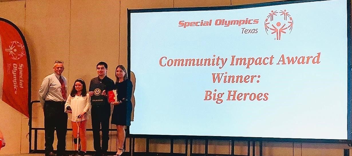 A group of people standing in front of a large screen that says community impact award winners big heroes