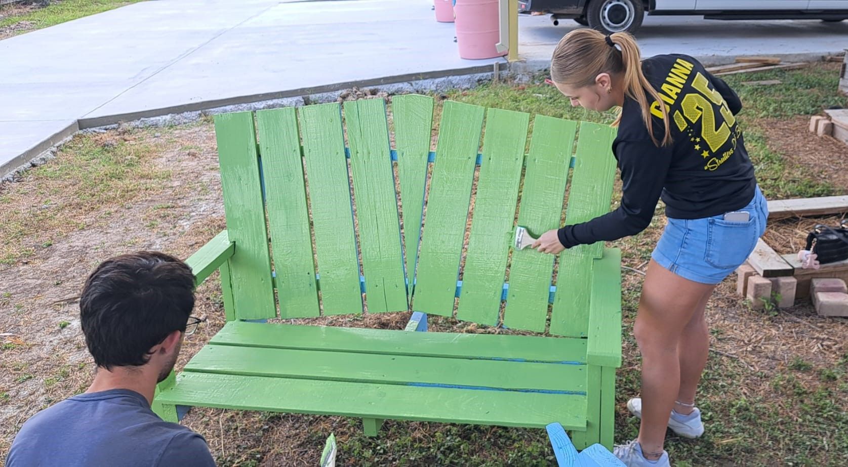 A man and a woman are painting a green bench.