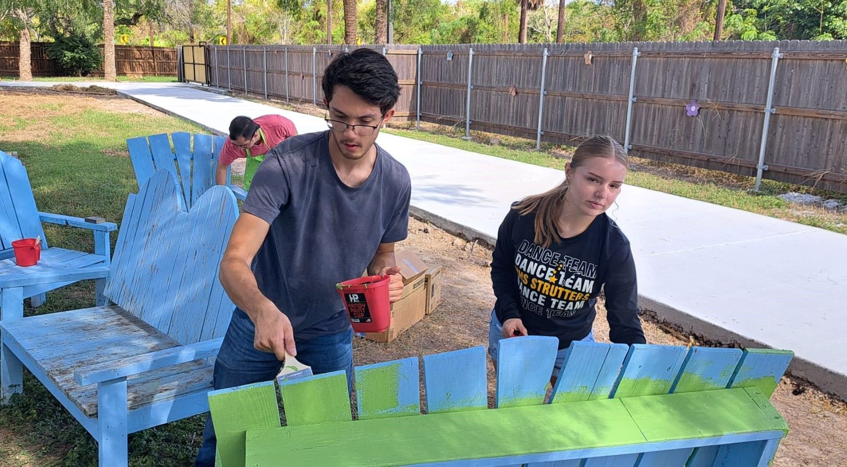 A man and a woman are painting a blue and green bench.