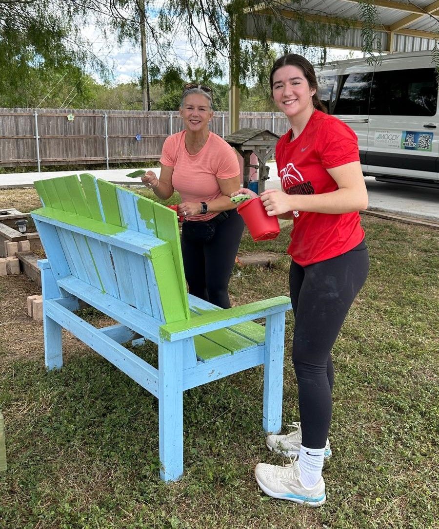 Two women are painting a blue and green bench in a yard.
