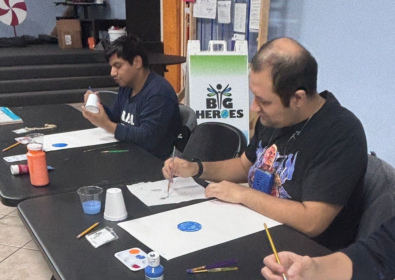 Three men are sitting at a table painting with a big heroes sign in the background.