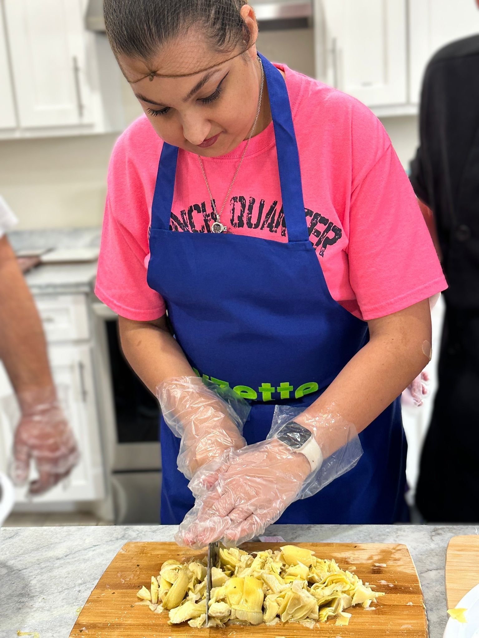 A woman in a pink shirt and blue apron is cutting artichokes on a cutting board