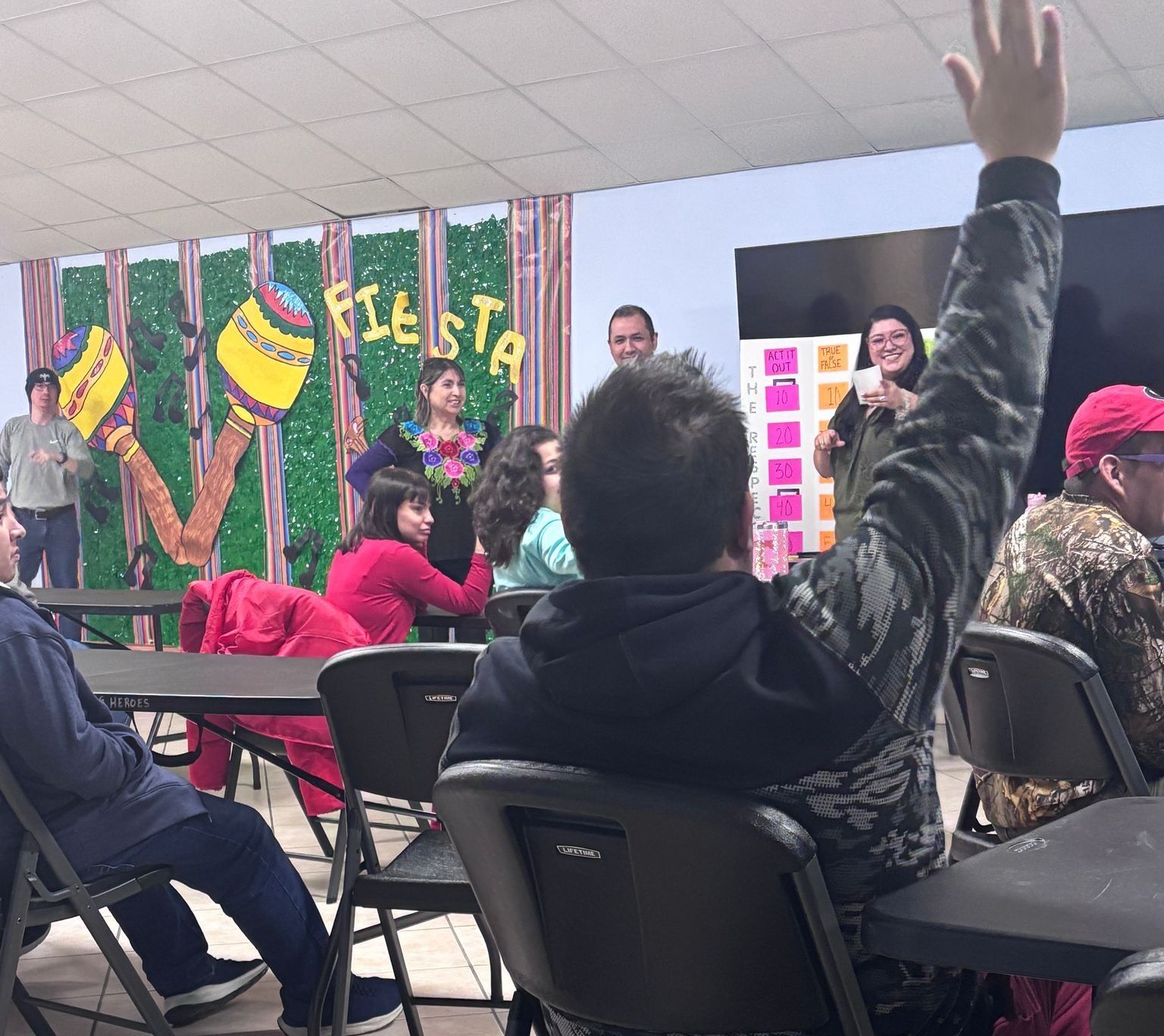 A group of people are sitting at tables with their hands up in front of a wall that says fiesta