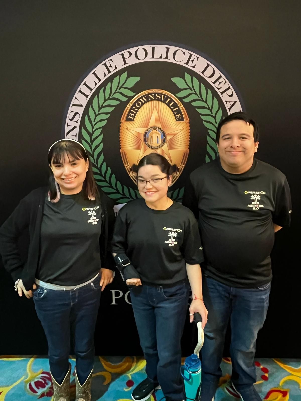 Three people stand in front of a Brownsville Police Department emblem. They wear black shirts and jeans.