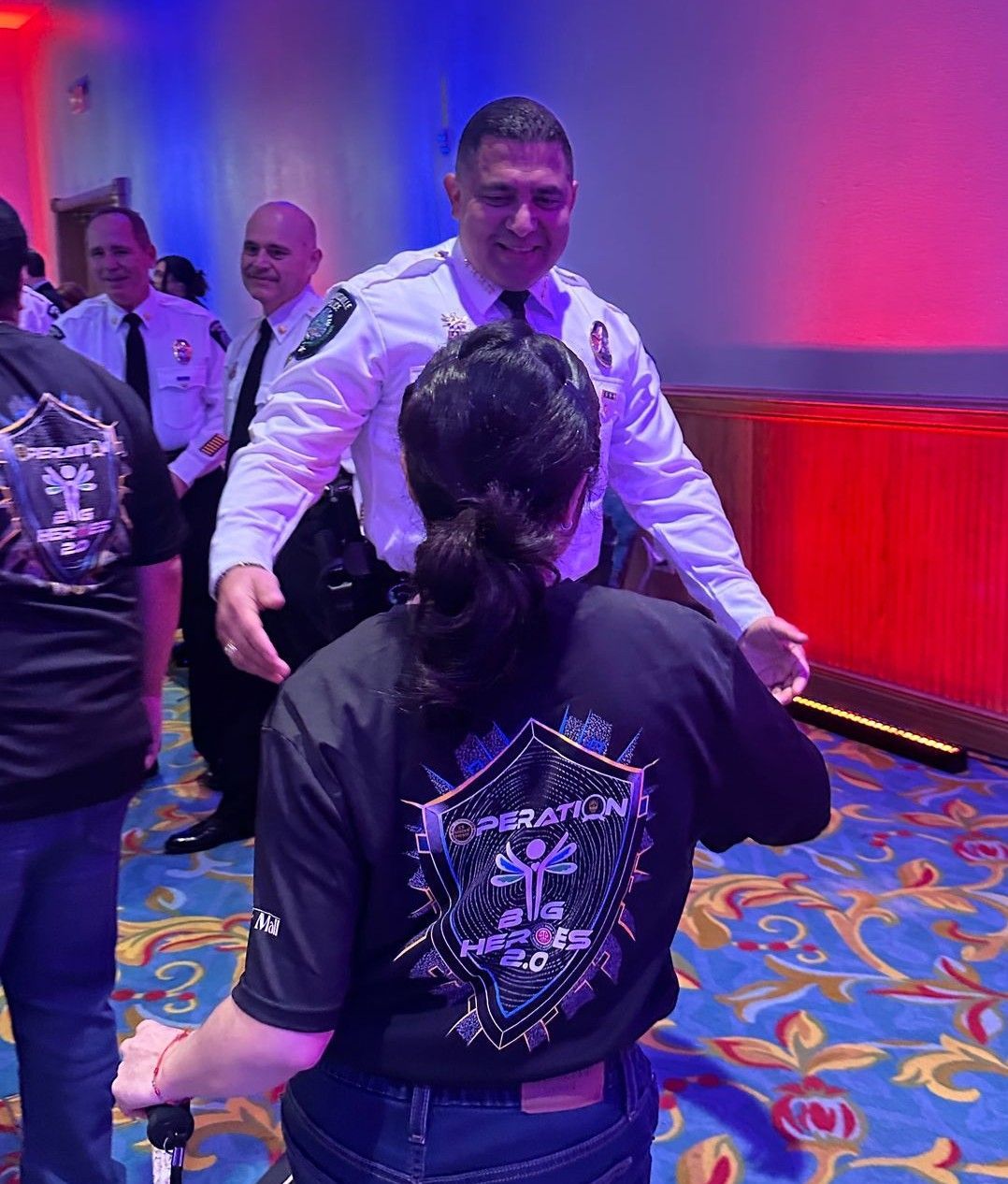 Man in uniform greets person wearing a shirt with “Inspiration” on the back, inside a brightly lit event.