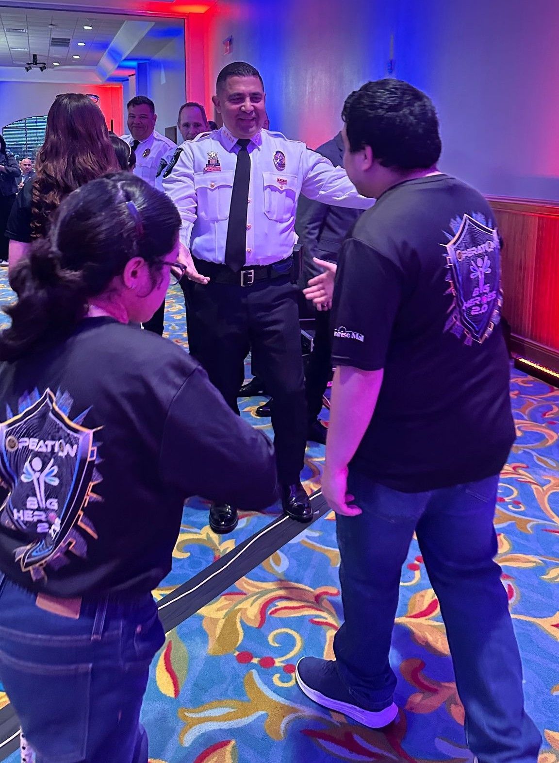 People in black shirts with logo greeting a man in a police uniform in a brightly lit room.