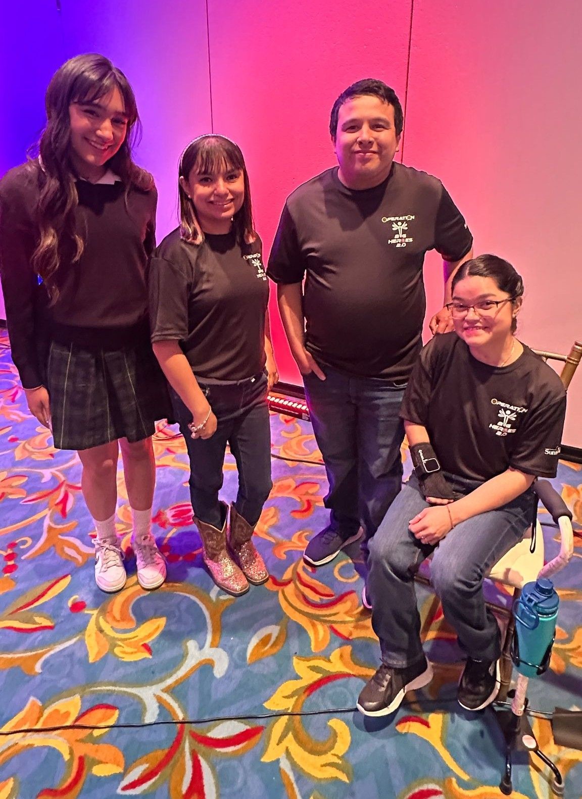 Four people stand on a stage with a floral carpet. Two girls wear school uniforms. Two wear black t-shirts. All smile.