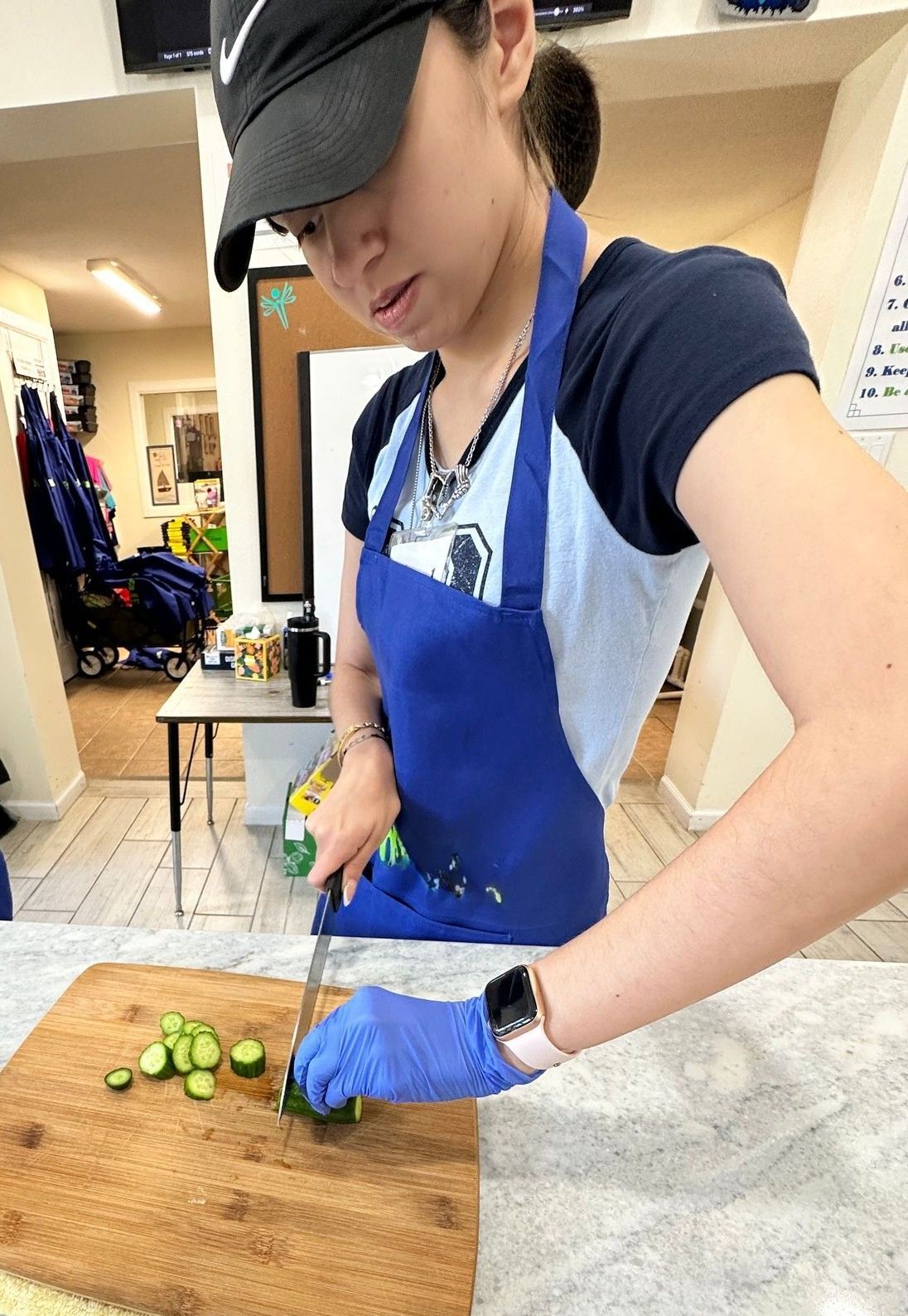 A woman in a blue apron is cutting cucumbers on a cutting board.