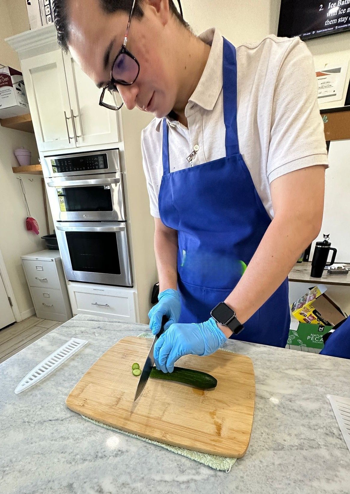 A man in a blue apron is cutting a cucumber on a cutting board.