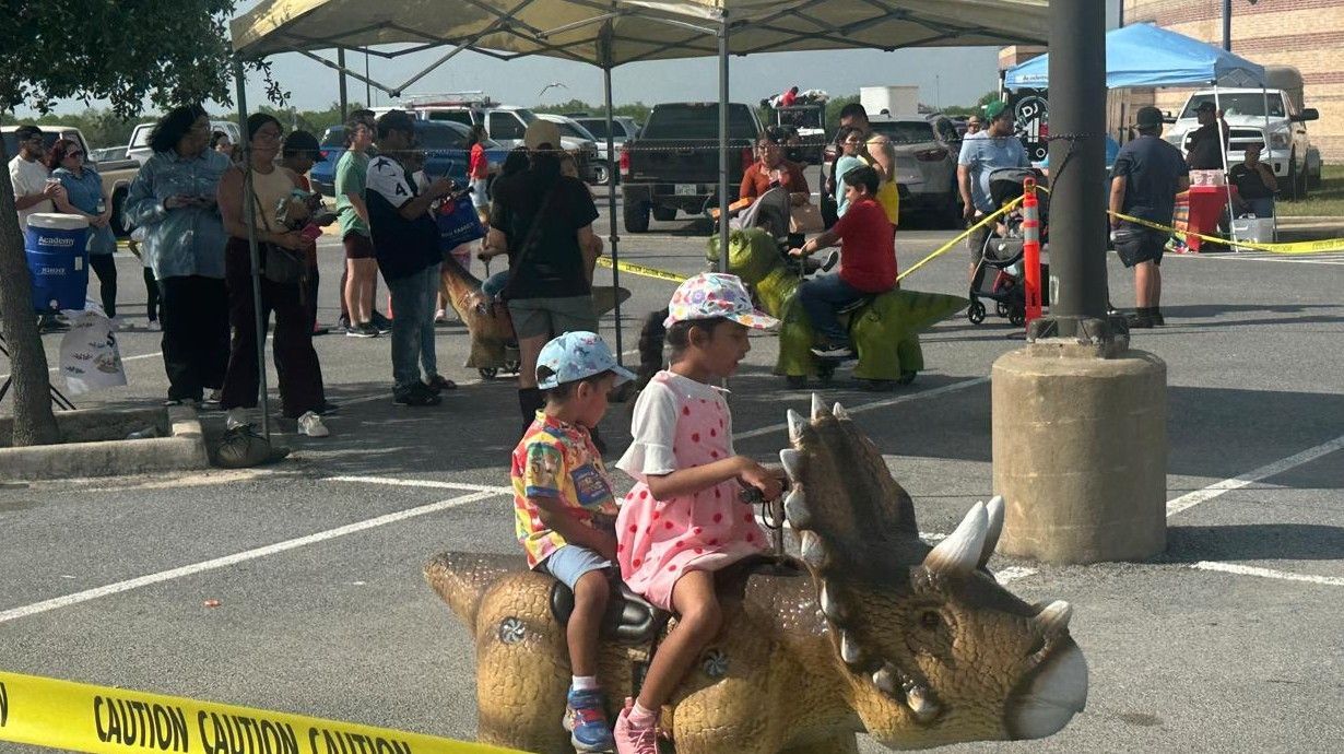 Two children are riding on a dinosaur statue in a parking lot.