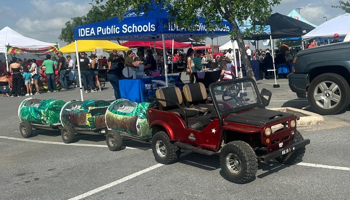 A red jeep is parked in a parking lot next to a blue tent that says idea public schools.