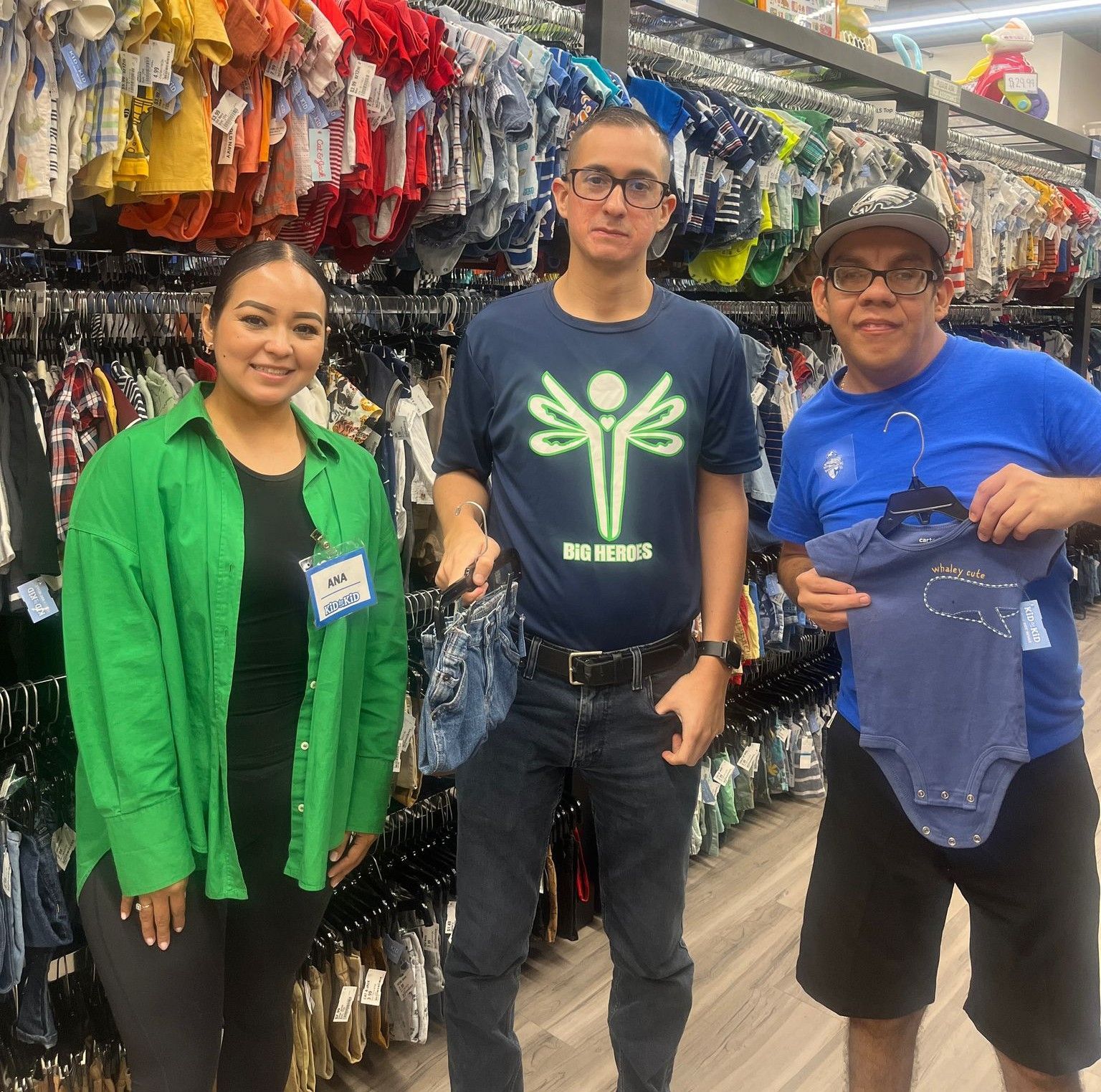 Three people in a thrift store. Two hold baby clothes. One woman wears green, two men wear blue.