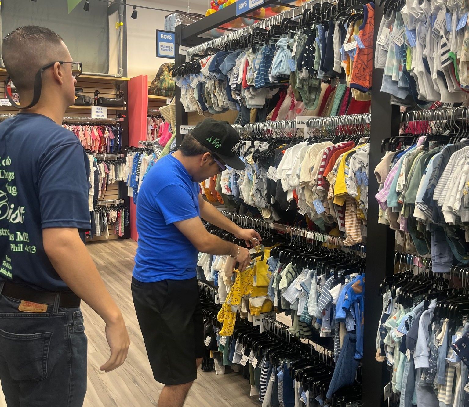 Two men shop for baby clothes in a store. One man examines a yellow outfit.