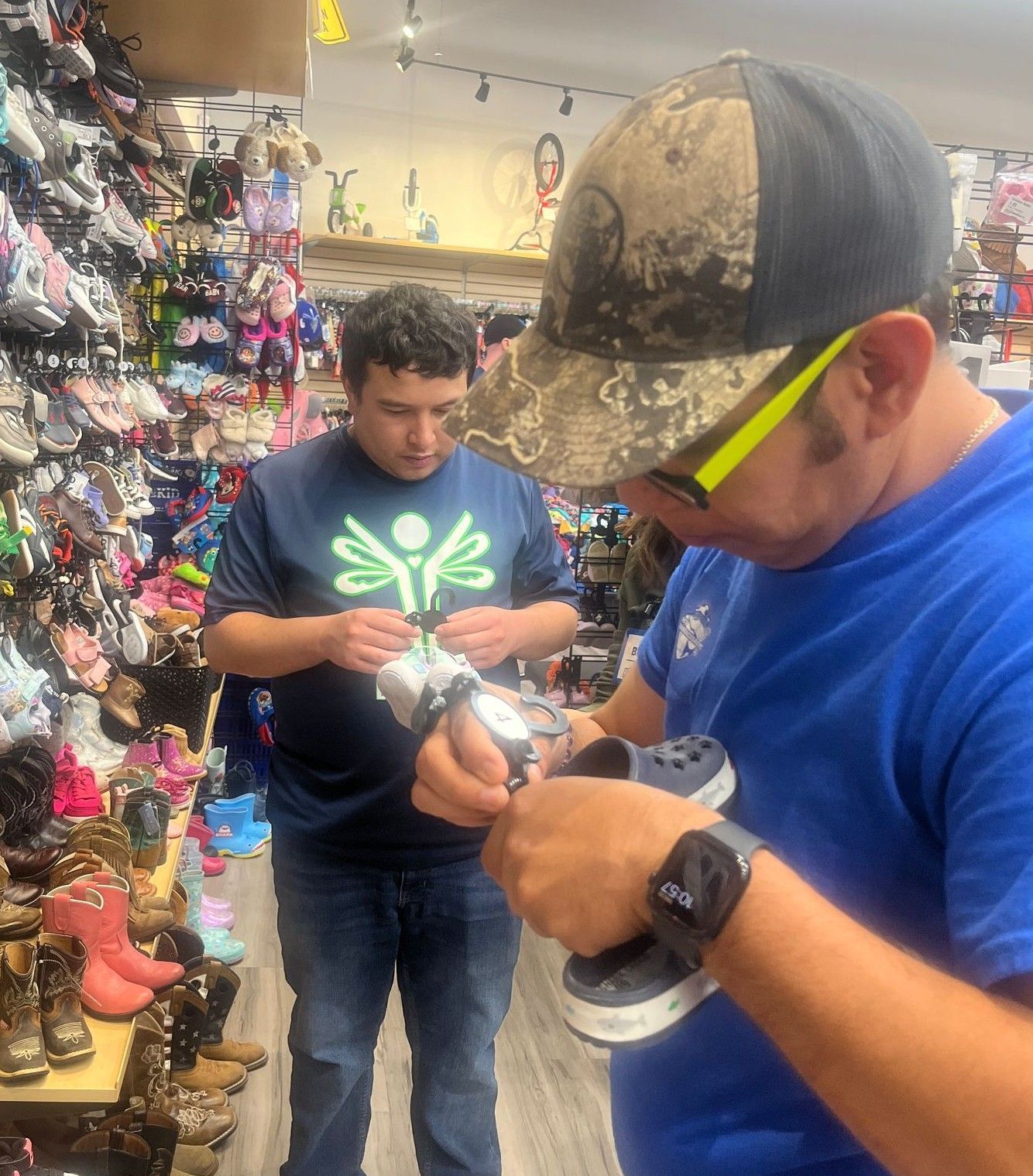 Two men in a shoe store look at Crocs. One wears a camo hat. Shoes line the walls.