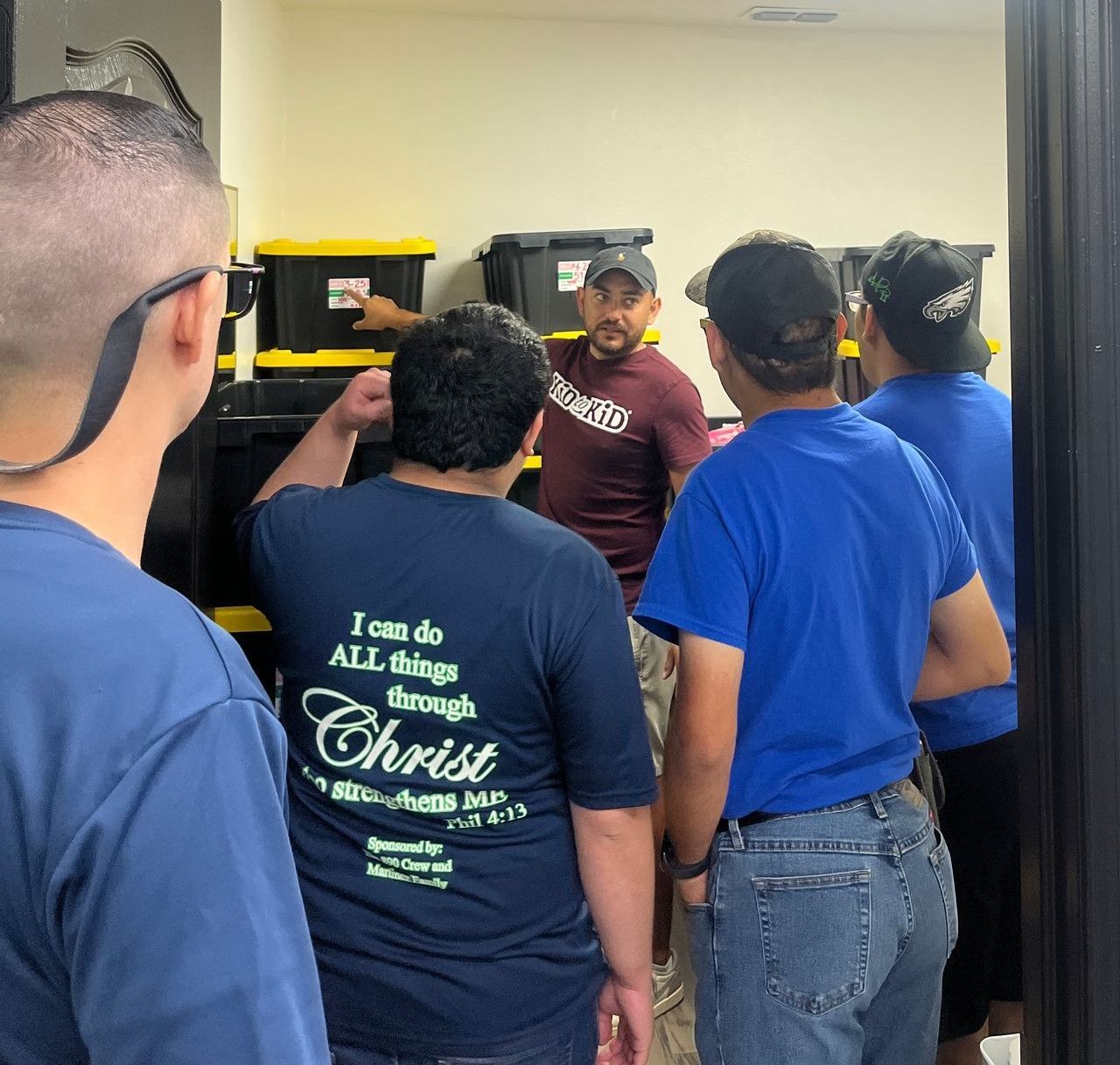 Men in room: a man in maroon shirt points to boxes, others listen.