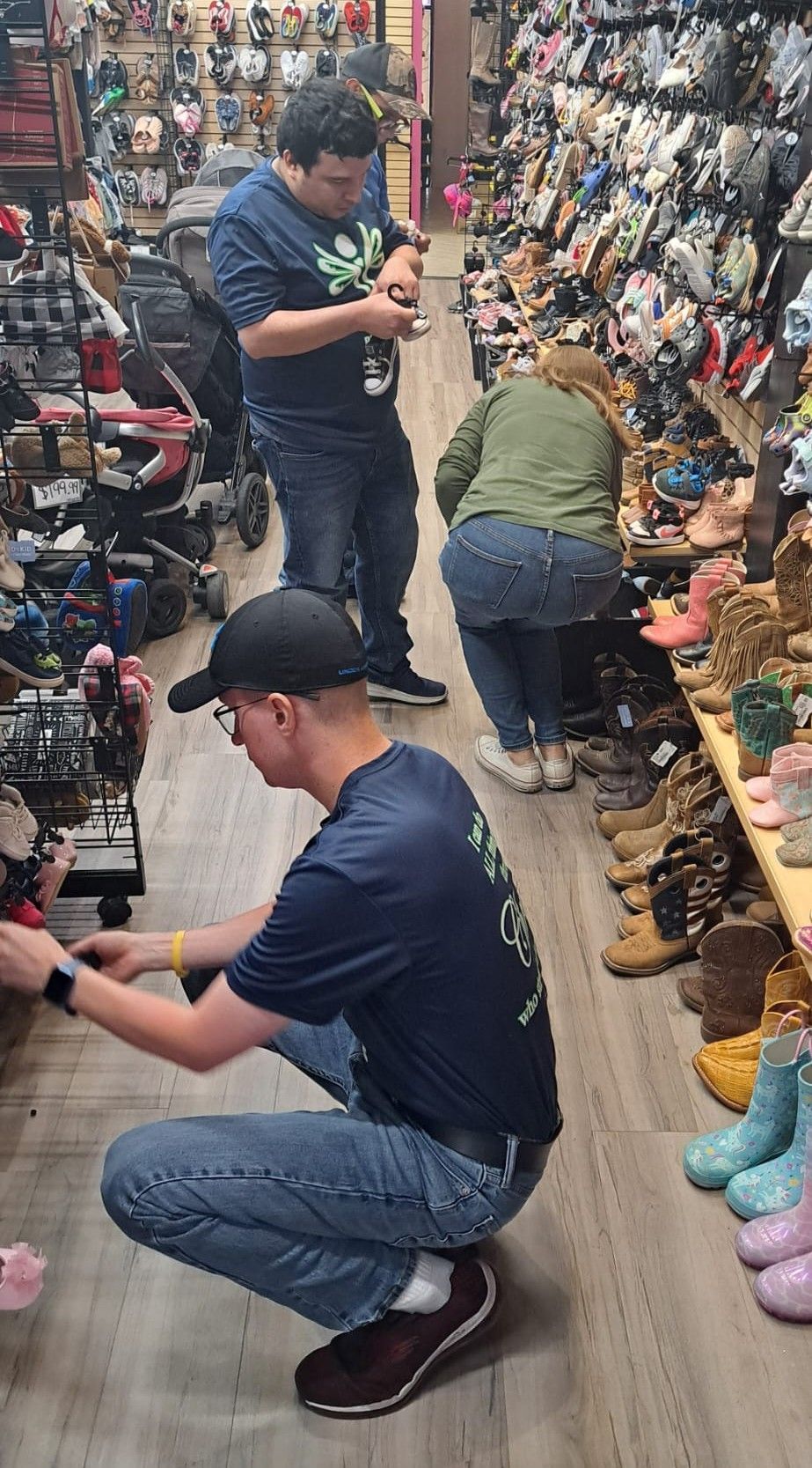 People browsing and inspecting shoes in a crowded shoe store. One person squats, another stands and leans.