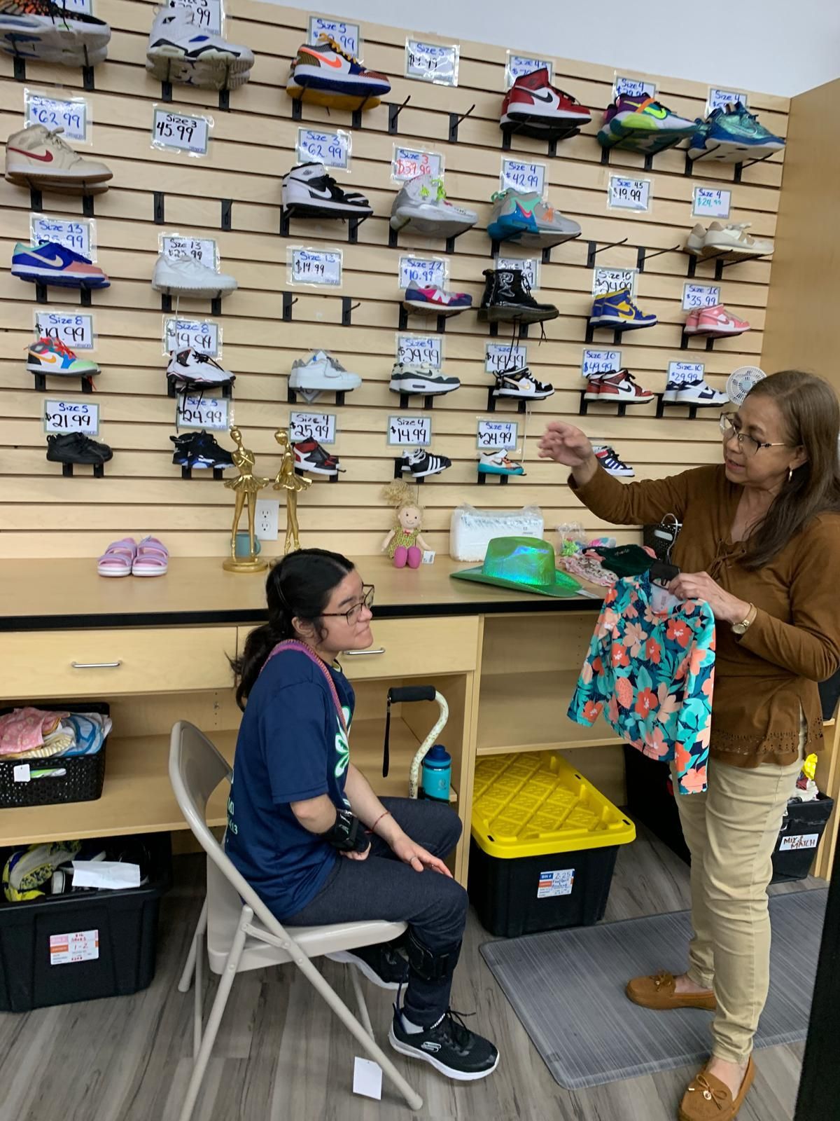 Woman shows dress to seated person in sneaker shop. Shoes line wall.