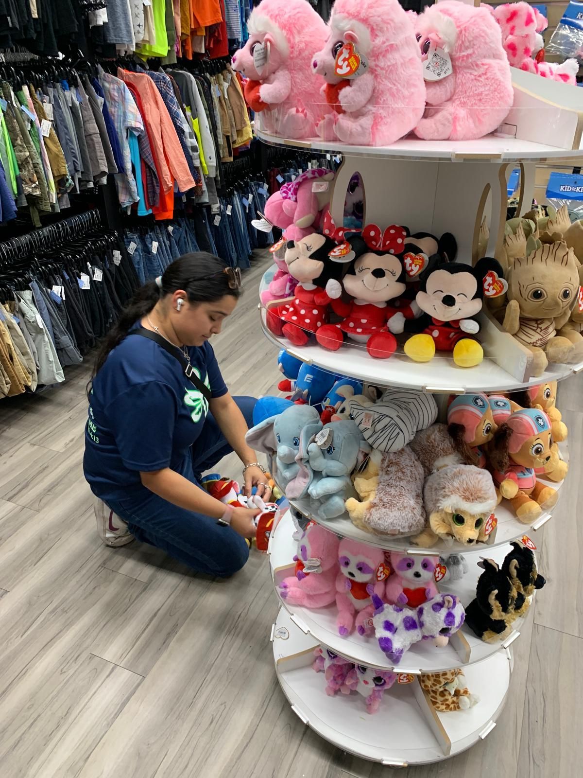 Woman arranging plush toys on a multi-tiered shelf in a store, clothing racks in the background.